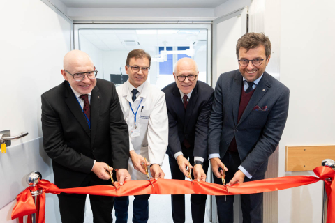 Four people stand in the doorway and simultaneously cut a red ribbon with scissors, symbolizing the grand opening of the room. In the background, a white door leads to a bright room with medical equipment.