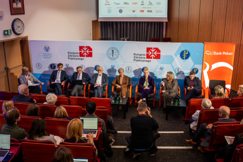A group of people sitting in the middle of the auditorium. They are participants in a panel discussion. Behind them is a wall with various logos. Participants in the event can also be seen.