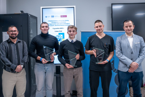 Pięć osób stoi obok siebie w pomieszczeniu. Każda osoba trzyma szklaną statuetkę w dłoniach. W tle widać ekran z tekstem dotyczącym olimpiady wiedzy i jej finału. 
