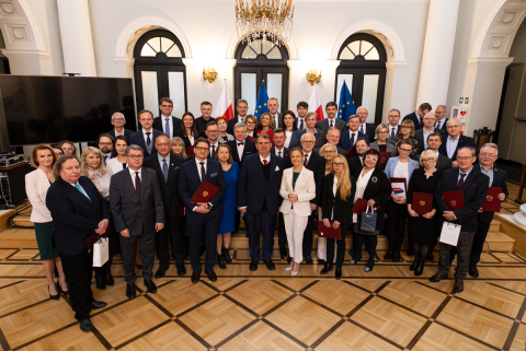 A group of people in an elegant room with parquet flooring and a chandelier, standing in front of the flags of Poland and the European Union. Some of them are holding burgundy briefcases with the Polish coat of arms.