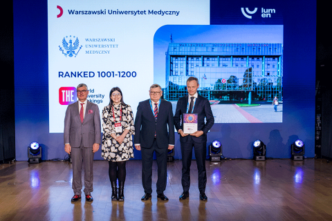 Four people are standing on stage in front of a large screen displaying the university's logo and ranking information. One person is holding a framed award. Everyone is looking at the camera.