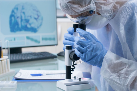 Naukowiec w maseczce czepku i fartuchu patrzy przez mikroskop