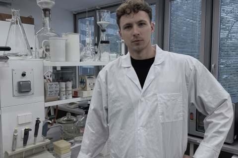 Osoba w białym fartuchu laboratoryjnym stojąca w nowoczesnym laboratorium chemicznym, otoczona kolbami, cylindrami miarowymi i innym sprzętem badawczym, na tle półek i dużych okien.