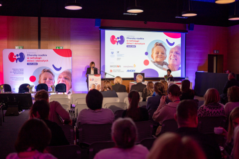 Kobieta przemawia z mównicy do zgromadzonej publiczności. Uczestnicy siedzą w rzędach i słuchają wystąpienia. Za prelegentką oraz na dużym ekranie widać grafiki i informacje o tematyce chorób rzadkich w nefrologii dzieci i dorosłych.