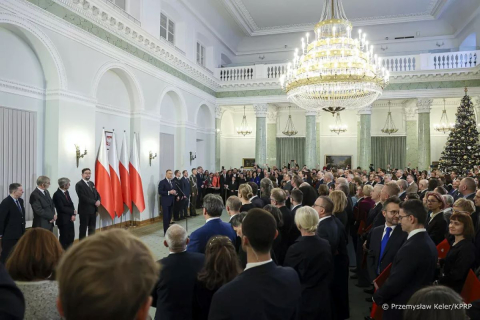 „Uroczystość w reprezentacyjnej sali z dużym żyrandolem i choinką, z udziałem licznych osób stojących w rzędach. Na pierwszym planie widoczna grupa gości, w tle przemówienie przy trzech flagach Polski ustawionych obok ściany. Przemawia prezydent RP.