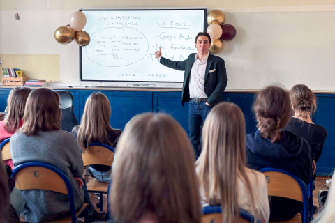 Grupa kilkunastu osób siedzi w sali i patrzy na osobę prowadzącą zajęcia stojącą przy tablicy. Prowadzący wskazuje ręką zapis na tablicy i stoi obok dużego ekranu z notatkami. W tle znajdują się dekoracje z balonów oraz półka z książkami. 