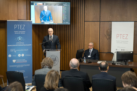 Po prawej stronie prelegent, który stoi za mównicą i przemawia. Obok prelegenta stoi roll‑up z informacjami o konferencji. Nad nim wisi ekran z transmisją wystąpienia. Po lewej - siedzi jeszcze jeden mężczyzna.