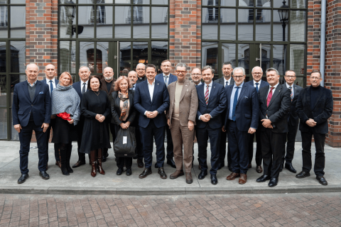 19 osób ustawionych w dwóch rzędach przed budynkiem z cegły i dużymi oknami. Osoby stoją blisko siebie i patrzą w stronę aparatu. Mają na sobie stroje formalne. Tło tworzą wysokie okna i elewacja z cegły. Grupę otacza brukowany chodnik.