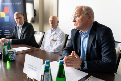 Three individuals are seated at a conference table with documents and water bottles in front of them, with an informational display visible in the background.
