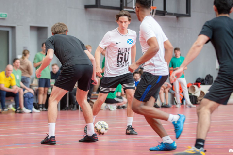 Players participating in an indoor football match are fighting for the ball in the center of the pitch, while the remaining participants and spectators watch the game from the sidelines.
