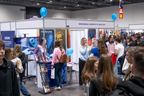 The trade hall features a booth of the Medical University of Warsaw, decorated in blue and displaying informational materials. Several people are looking at brochures and talking at the booth. In the background, other stands and visitors can be seen.