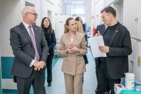 A group of people is talking during a visit in a hospital corridor; one of them is holding documents, while the others listen and stand nearby in formal attire.