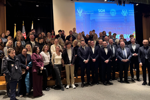 Around fifty people are standing and sitting on the steps of the stage for a group photo. In the front row, a group of men in suits are standing and looking toward the photographer. Behind them, more people are standing, dressed both elegantly and casually. Several people are raising their hands in a gesture of joy. In the background, there is a large screen displaying the university’s logos.
