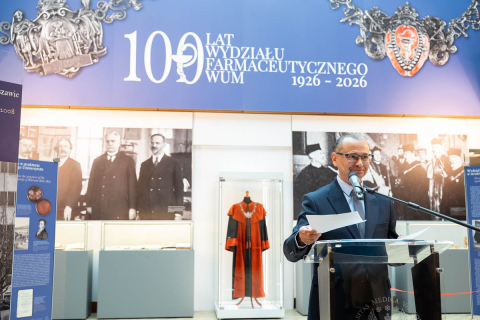 A speaker delivering an address during the ceremony marking the 100th anniversary of the WUM Faculty of Pharmacy, set against a backdrop of historical displays and showcases with university memorabilia.