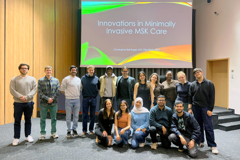 Grupa kilkunastu osób pozuje do zdjęcia. W tle widać duży ekran multimedialnej. Na ekranie wyświetlony jest slajd z tytułem „Innovations in Minimally Invasive MSK Care” oraz nazwiskiem prowadzącego. Uczestnicy ustawieni są w dwóch rzędach – część stoi, a część siedzi na podłodze z przodu.