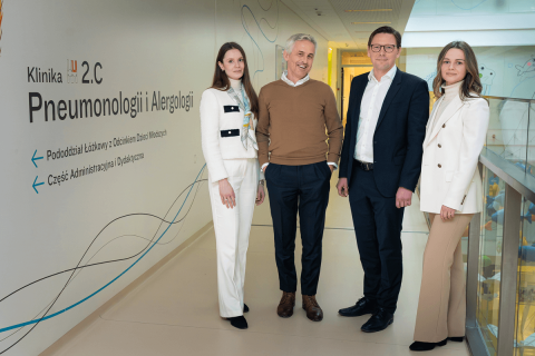 Two women and two men, all elegantly dressed, pose for a photo in a hospital corridor. The name of the clinic and several colorful lines can be seen on the corridor wall.