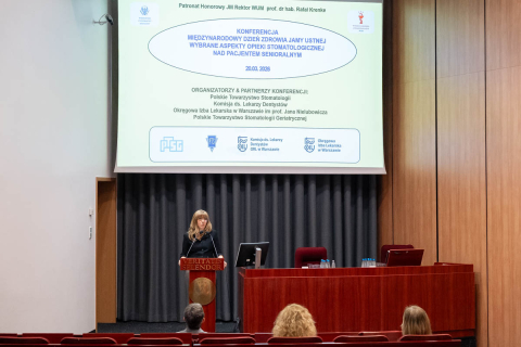 Osoba stojąca przy mównicy na tle dużego ekranu z prezentacją o immunosenescencji. Osoba ma jasne włosy, jasnoniebieską marynarkę i trzyma pilot do slajdów. Przed mównicą siedzą dwie osoby zwrócone w stronę sceny.