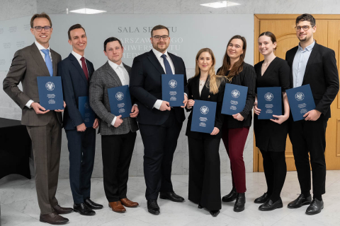 A group of young, elegantly dressed people poses for a photo, holding navy-blue folders in their hands