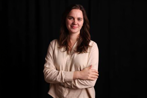 Osoba stojąca na tle ciemnego jednolitego tła, ubrana w jasną bluzkę z długim rękawem i skrzyżowanymi przedramionami.