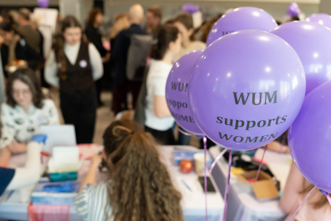 Stoisko podczas wydarzenia wspierającego kobiety, udekorowane fioletowymi balonami z napisem ‘WUM supports WOMEN’. W tle widoczni uczestnicy zgromadzeni przy różnych stanowiskach.