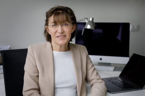 Profesor Hanna Szajewska siedzi na krześle przy biurku. Ma na sobie jasną marynarkę i białą bluzkę. W tle znajduje się komputer stacjonarny z dużym monitorem, lampka biurkowa na metalowym wysięgniku oraz otwarty laptop.