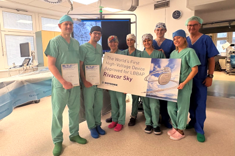 Zdjęcie przedstawia grupę ośmiu osób stojących w sali zabiegowej. Wszystkie osoby mają na sobie odzież medyczną: czepki, maseczki opuszczone pod brodę, fartuchy i ochraniacze na obuwie. Dwie osoby po lewej stronie trzymają dyplomy. Kilka osób po prawej stronie trzyma duży prostokątny baner. Na banerze znajduje się tekst: ‘The World's First High-Voltage Device Approved for LBBAP – Rivacor Sky’ oraz logo firmy Biotronik. W tle widać monitor z obrazem medycznym, lampy operacyjne i sprzęt zabiegowy.