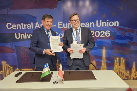 Dwie osoby stoją za stołem konferencyjnym i trzymają w dłoniach podpisane dokumenty. Na stole ustawione są małe flagi Polski i Uzbekistanu oraz teczki na dokumenty. W tle znajduje się baner z napisem „Central Asia–European Union University Congress 2026”