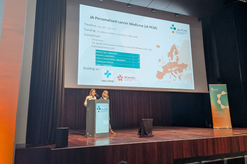 Two presenters stand at a podium on a conference stage, delivering a presentation about the “JA Personalized Cancer Medicine (JA PCM)” initiative, with a slide showing a map of Europe, consortium details, and partner logos.
