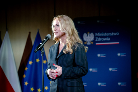 A woman standing at a microphone, dressed in a dark suit. The woman has her hands clasped at stomach level. In the background there are the flags of Poland and the European Union and a navy-blue backdrop with the coat of arms and the inscription “Ministry of Health”.