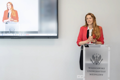 A person stands behind a transparent lectern bearing the emblem of the Medical University of Warsaw and delivers a speech. A large presentation screen stands nearby, displaying an image of the same person speaking at the lectern. The interior of the room is bright, with a light-colored wall in the background.