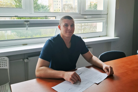 Młody chłopak siedzący za biurkiem, na którym widać kartki papieru. W tle, za nim, są okna.