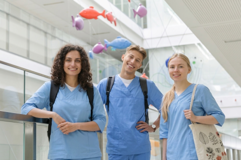 Trzy młode osoby ubrane w niebieskie fartuchy medyczne pozują do zdjęcia w jasnym pomieszczeniu. W tle widać kolorowe rybki zwisające z dachu.