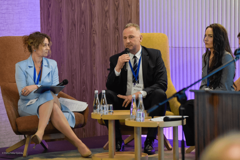 Trzy osoby siedzą na scenie w czasie konferencji. Są elegancko ubrane. Jedna z nich, mężczyzna, prof. Sobczak, trzyma w rękach mikrofon i przemawia.