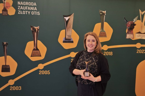 Osoba stojąca na tle zielonej ścianki ekspozycyjnej i trzymająca w dłoniach statuetkę nagrody; w tle widoczne różne statuetki oraz napis „Nagroda Zaufania Złoty Otis” wraz z graficzną osią czasu z zaznaczonymi latami.