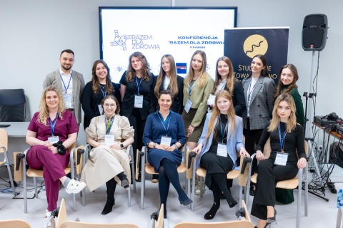 Grupa kobiet i mężczyzn pozuje do wspólnego zdjęcia w sali konferencyjnej. Część kobiet i mężczyzn siedzi na krzesłach w pierwszym rzędzie, a pozostali stoją za nimi. Wszyscy mają identyfikatory na smyczach, a osoby siedzące w pierwszym rzędzie trzymają w rękach aparaty fotograficzne lub mają dłonie ułożone na kolanach. W tle znajduje się ekran z napisem „Konferencja Razem dla Zdrowia” oraz baner Studenckiego Towarzystwa Naukowego.