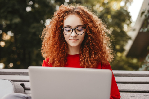 Młoda dziewczyna w okularach siedzi na ławce w parku i patrzy w ekran rozłożonego laptopa.