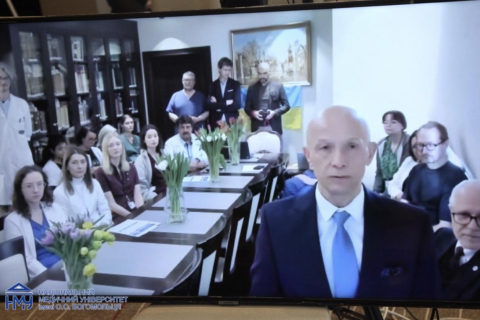 A conference room with a long table and several people seated on both sides. At the front, one person in a suit stands facing the camera. On the table there are vases with tulip flowers and printed documents. Bookshelves and a framed picture are in the background.