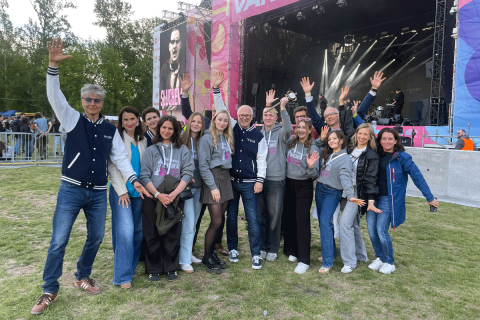 A group of people posing together outdoors at a music festival, standing on grass near a large concert stage. Several people are raising their hands, and a performance stage with screens and lighting is visible in the background.