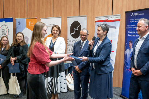 A formal award ceremony taking place in a lecture hall, where a young attendee in a red blouse and dark skirt receives a diploma from a speaker holding a microphone and a document. Several other attendees stand in a row behind, holding folders and tote bags, with informational banners displayed in the background.