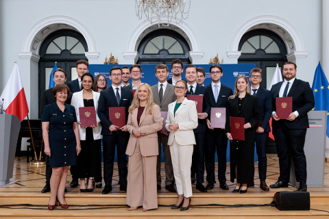 A large group of people stands on steps inside an elegant hall, holding red diploma folders with a national emblem. In front of the group stand several individuals posing for a formal photo. Polish and European Union flags are placed on both sides, and a blue backdrop with the text “Ministerstwo Zdrowia” is visible behind them.