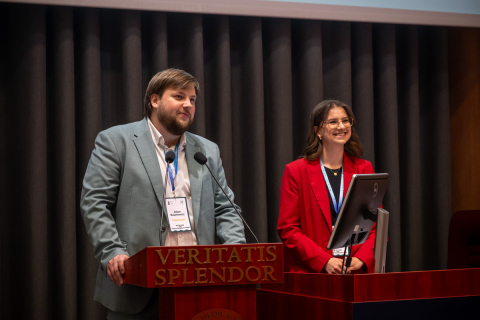 Dwóch prelegentów stoi przy mównicy z napisem „Veritatis Splendor” w sali wykładowej. Mężczyzna w jasnej marynarce mówi do mikrofonów i trzyma dłonie oparte na mównicy, a kobieta w czerwonej marynarce stoi obok pulpitu z monitorem komputerowym i ma przypięty identyfikator na smyczy.