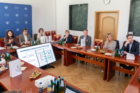 Kobiety i mężczyźni biorą udział w dyskusji przy stole konferencyjnym w sali Warszawskiego Uniwersytetu Medycznego. Część uczestników trzyma długopisy i notatniki, a na ekranie ustawionym na stole prezentowana jest plansza dotycząca architektury rozumowaniaklinicznego.