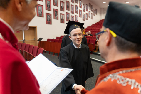 A person in a gown and mortarboard is shaking hands with another person dressed in formal attire during a ceremony in a hall with red seats. On the left side of the frame, another person is holding an open diploma. In the background, framed portraits hang on the wall.