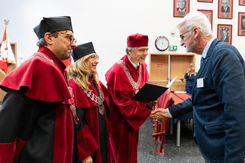 Three people and one additional person dressed in red and black academic gowns and caps stand in a lecture hall. Two people and one additional person wear decorative rector’s chains. One person in a red cap holds an open folder with a document and shakes hands with another person in a suit. In the background, there are red chairs, a door, and a wall with framed photographs.