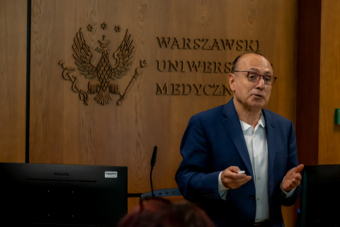 A man stands in a lecture hall at Warsaw Medical University and speaks during a presentation, holding a remote control in his hand