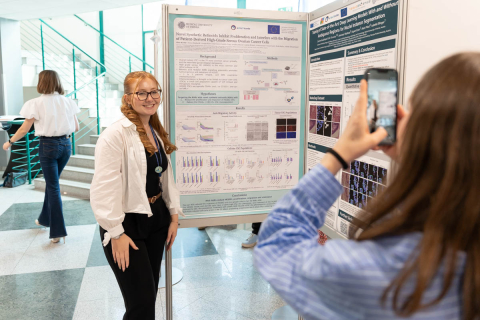 One person stands next to a scientific poster presentation board and poses for a photo. Another person in the foreground holds a smartphone and takes the picture. The poster contains charts, diagrams, and scientific text. In the background, a staircase and another person walking are visible.