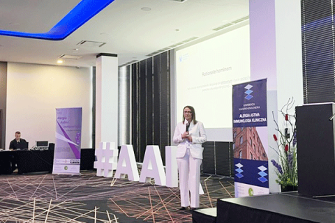 Kobieta stoi na środku sali konferencyjnej i przemawia. Po lewej duży napis „#AAI”. W tle ekran z prezentacją i roll‑upy z napisem „Alergia Astma Immunologia Kliniczna”. 