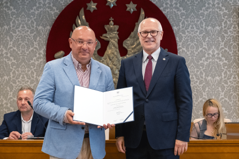 Dwóch mężczyzn pozujących do zdjęcia. Mężczyzna po lewej trzyma otwarty dyplom. Na drugim planie widać dwie siedzące osoby oraz duże godło WUM - złoty orzeł na czerwonym tle.