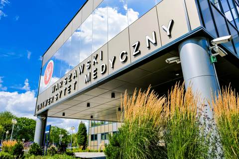 Budynek z przeszkloną fasadą. Na fasadzie litery tworzące napis WARSZAWSKI UNIWERSYTET MEDYCZNY oraz czerwono-białe godło WUM o okrągłym kształcie. Wokół budynku roślinność. 