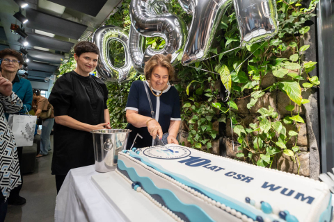 Dwie kobiety stoją przy stole i kroją tort jubileuszowy „10 lat CSR WUM”. Kobieta po prawej trzyma w dłoni nóż i kroi tort, a kobieta po lewej stoi obok. Obok stołu stoi metalowe wiadro. W tle wiszą srebrne balony oraz zielona ściana z roślin.